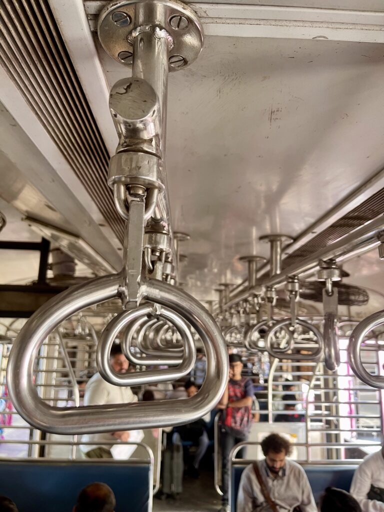 The inside of a compartment in Mumbai's suburban local trains. The focus is on the handles that people grab on to for support. There are a few people in the compartment and some seats are empty.